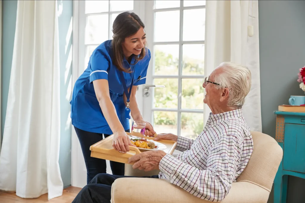 Caring in-home care nurse serving dinner to a senior man in an armchair in Montgomery County, Maryland.